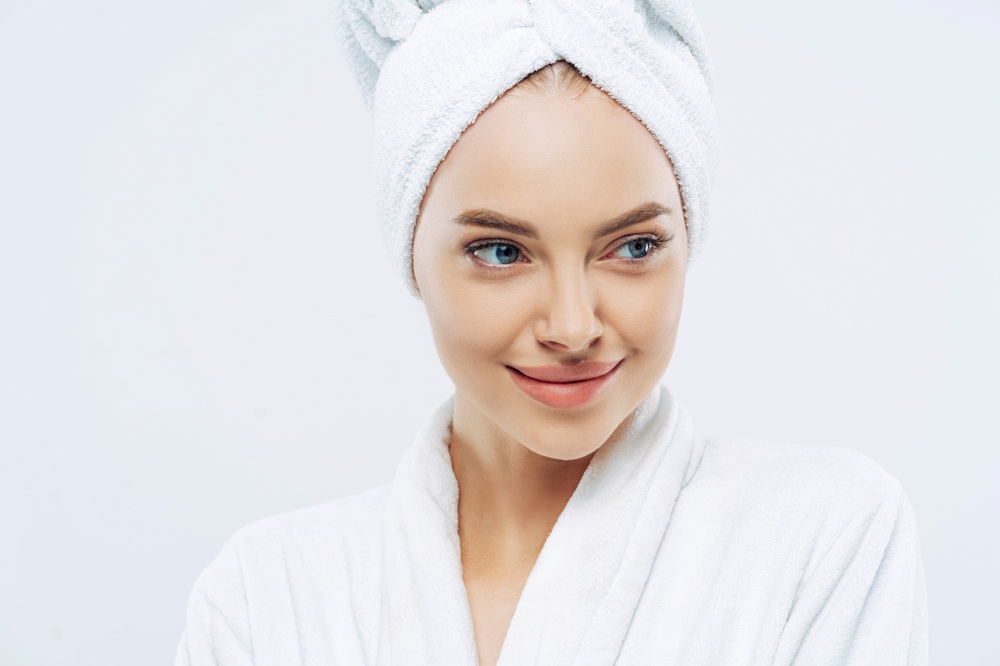 Pensive woman with healthy skin, refreshed after bath, dressed in gown, towel on head