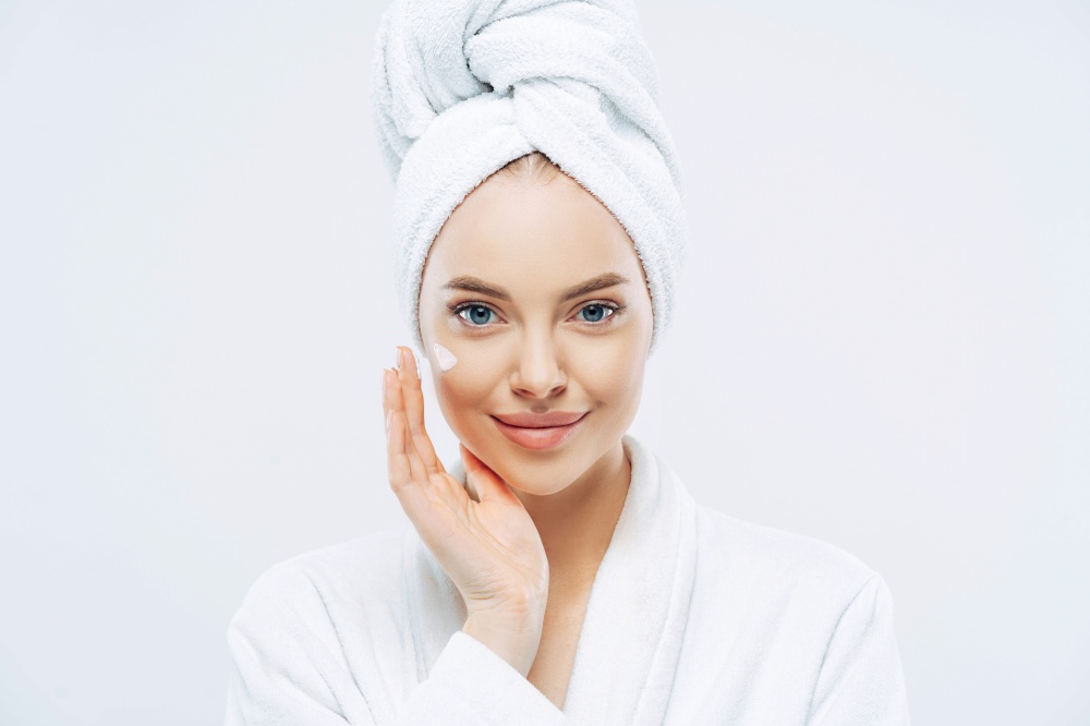 Confident woman applies anti-aging cream, enjoys new product, minimal makeup, bath robe, isolated.