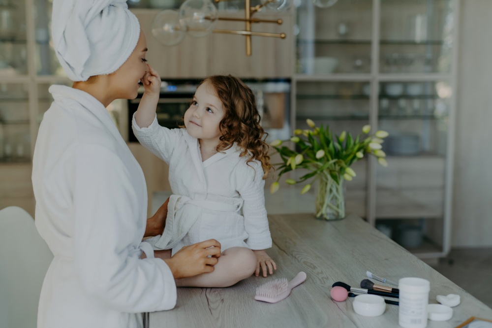 Curly girl touches mom&rsquo;s nose, in white robe, on cosmetic-filled table, preparing to do makeup. Cozy home interior.