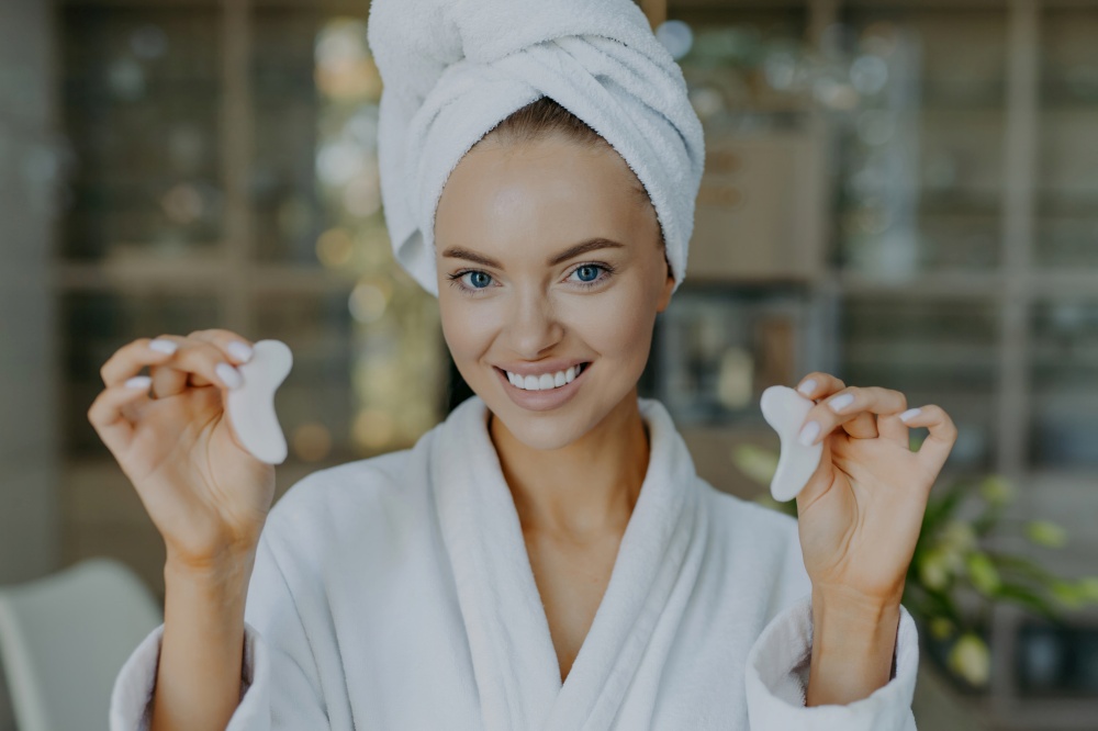 Smiling European woman with fresh skin holds patches, removes dark circles, wears bathrobe, positive gaze.
