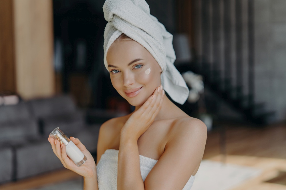 European woman applies face cream, hydrates skin. Minimal makeup, towel on head. Home interior, facial treatment. Anti-aging procedure.