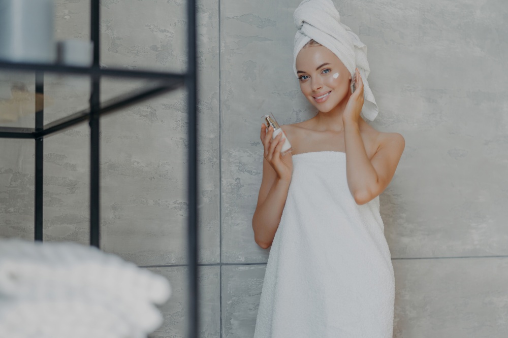 Happy woman with glowing skin applies cosmetic product, wrapped in towel, indoors. Bathroom background.