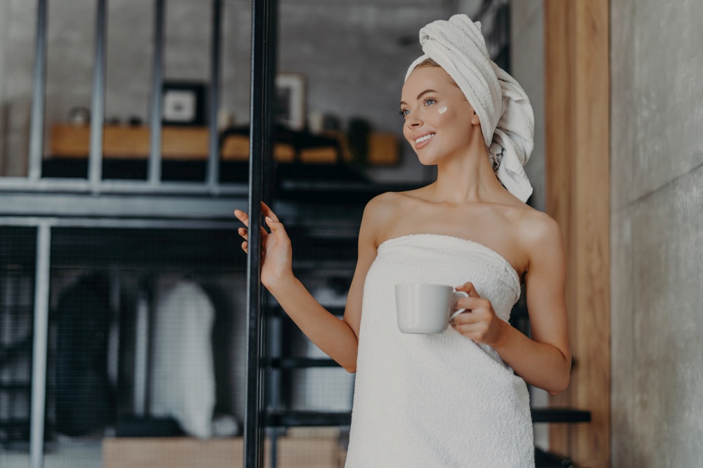 Thoughtful woman smiles, applies cream, wrapped in towel, drinks tea, relaxes on indoor stairs after bath