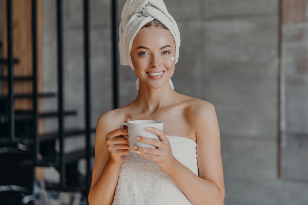 Smiling woman with makeup, toothy smile, bare shoulders, towel wrapped, drinks, applies moisturizing cream