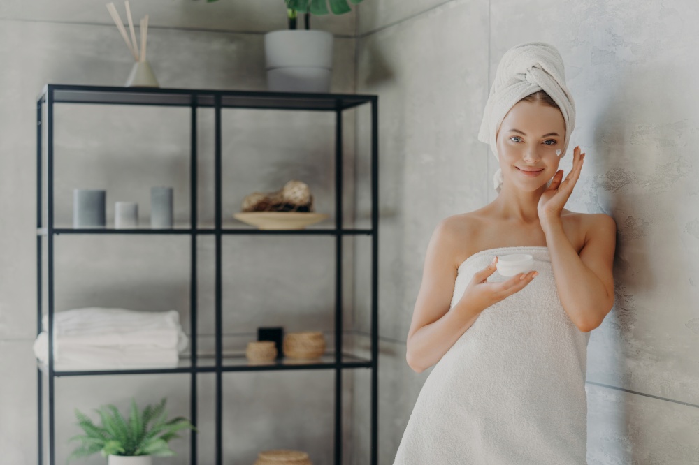 Healthy model uses cream, holds cosmetic jar, wrapped in towel, near cozy bathroom wall. Skin care