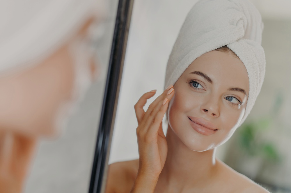 Woman gently touches face, admires reflection, enjoys soft skin, towel-wrapped head, minimal makeup. Beauty concept.
