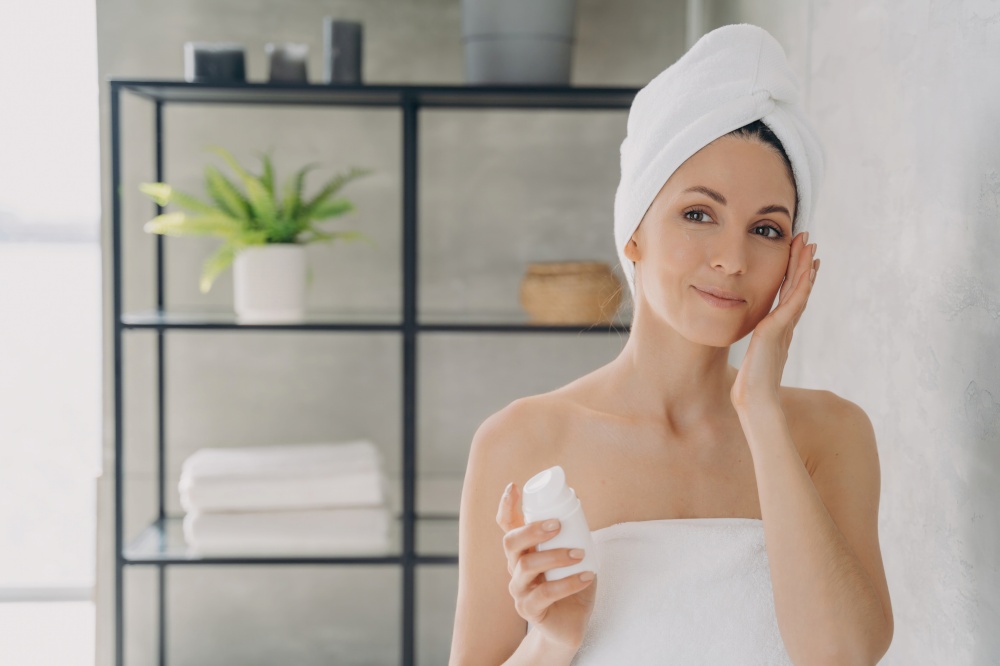 European lady applies face lotion, attractive girl with towel after bathing. Spa resort, face moisturizer, lifting cream.