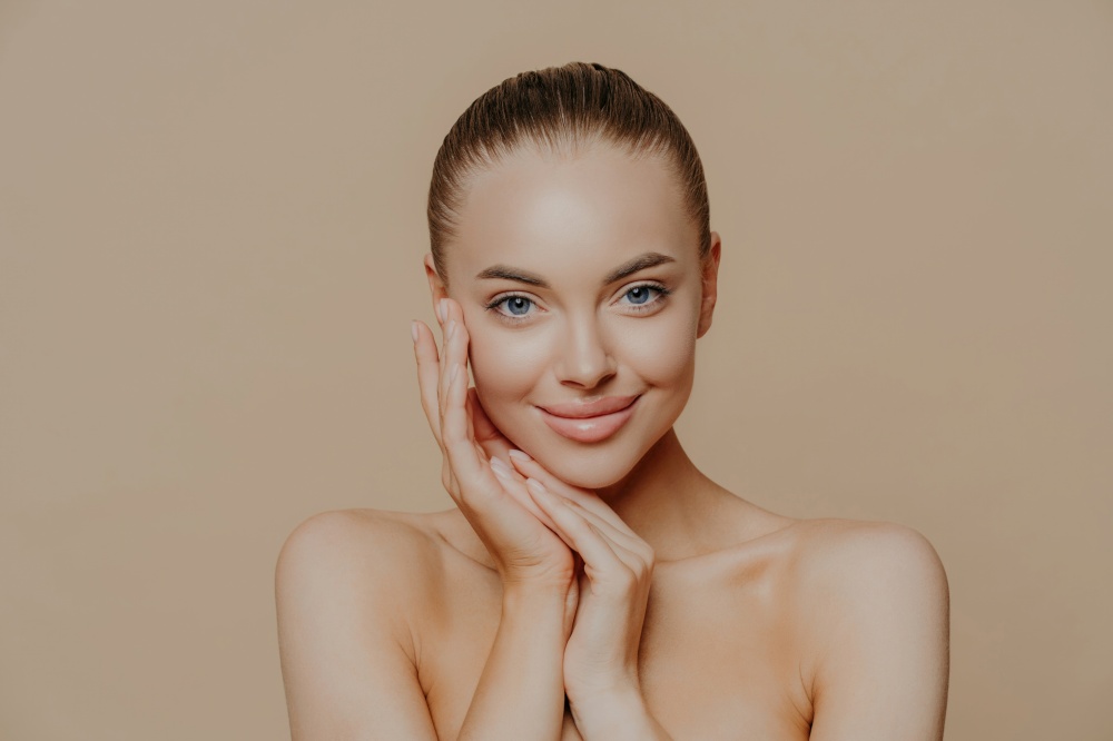 Natural beauty portrait. Fresh skin, gentle smile. Naked pose against beige background. Face touch, minimal makeup.