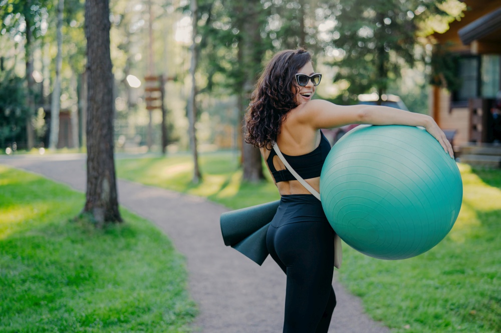 Happy brunette carries fitball and fitness mat in sport clothes, poses in green park. Good physical shape showcases healthy lifestyle and hobby.