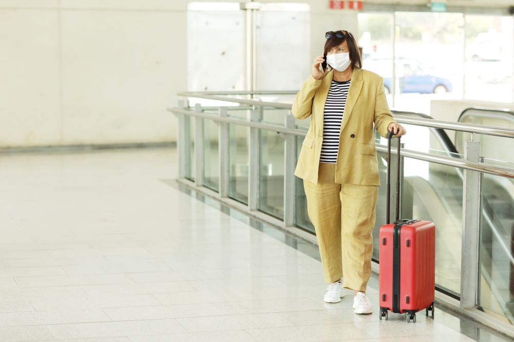 adult woman in protective antiviral mask is talking in smartphone in the airport. Woman in yellow suit with red suitcase waiting for the flight in protected sanitary mask. Copy space.. adult woman in protective antiviral mask is talking in smartphone in the airport. Woman in yellow suit with red suitcase waiting for the flight in protected sanitary mask. Copy space
