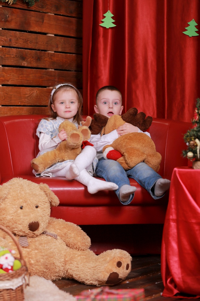 little girl and boy on sofa in christmas studio in red colors. christmas tree and teddy bear on background.. little girl and boy on sofa in christmas studio in red colors. christmas tree and teddy bear on background