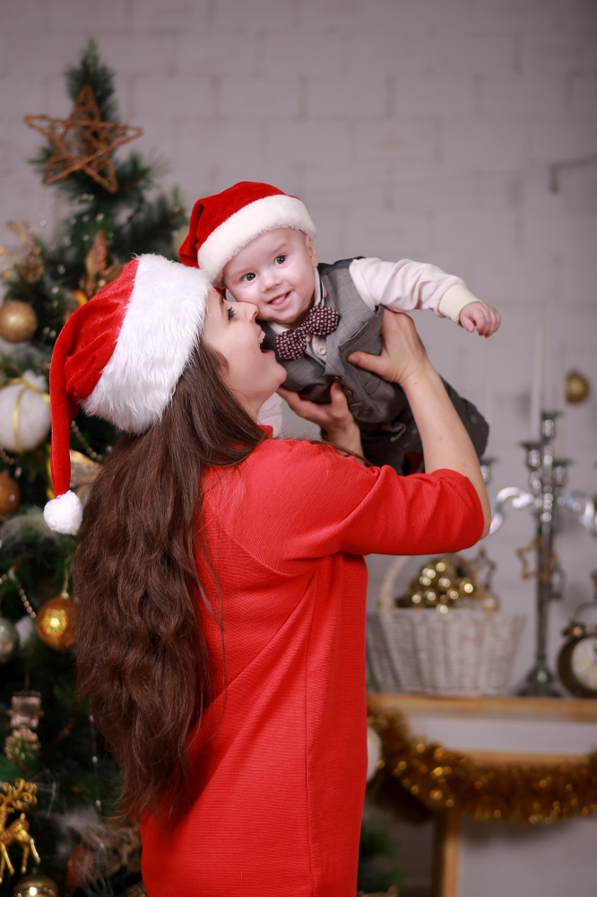 Young attractive happy mother have a good time with her son at home near christmas tree. Family, happiness, holidays, new year concept. Young attractive happy mother in santa&rsquo;s hat have a good time with her son at home near christmas tree. Family, happiness, holidays, new year concept