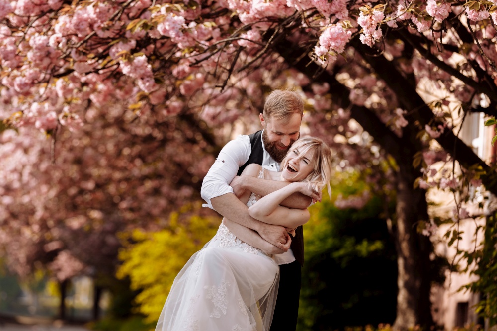 Beautiful, cheerful and lively newlyweds, groom and bride are hugging near the blooming pink cherry blossom. Wedding portrait of a close-up of a smiling bearded groom and a cute bride. Beautiful, cheerful and lively newlyweds, groom and bride are hugging near the blooming pink cherry blossom. Wedding portrait of a close-up of a smiling bearded groom and a cute bride.