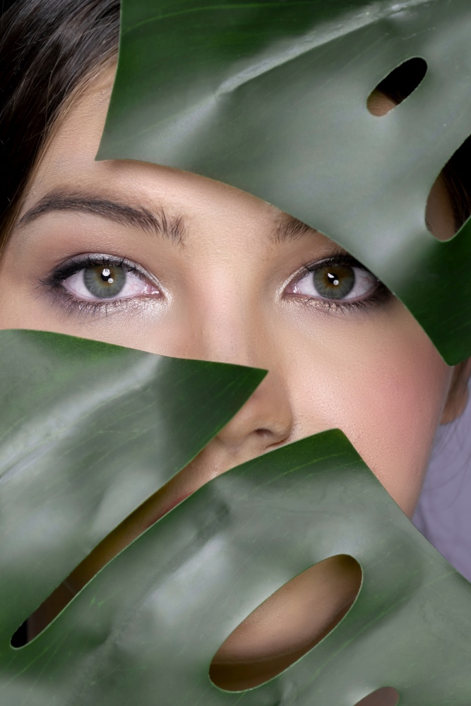 Portrait of young woman face surrounded with tropical green leaves. Healthy clean and perfect smooth skin of charming young woman. Natural beauty cosmetic and wellness concept for skincare product.. Closeup portrait face of charming girl covered with green leaf in beauty concept