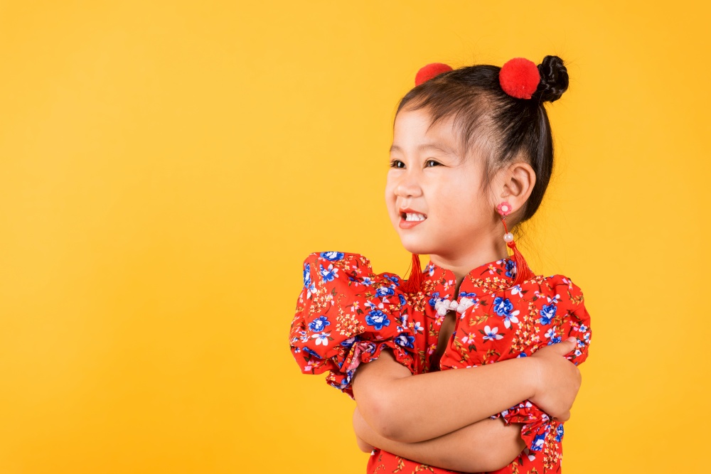 Happy Asian Chinese little girl smile wear red cheongsam crossed arms, Portrait children in traditional dress with greeting celebration for Chinese New Year, studio short isolated on yellow background