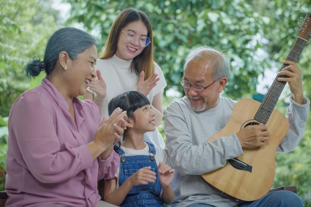 Happy family Asian senior elderly man or grandfather playing guitar while his grandmother and granddaughter singing song together outdoors at home, Activity family, Enjoying lifestyle senior older