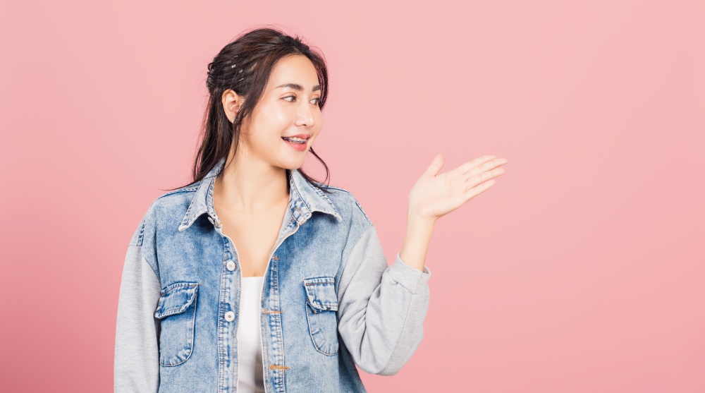 Asian happy portrait beautiful cute young woman smile standing presenting product holding something on palm away side, studio shot isolated on pink background with copy space, female show hand gesture