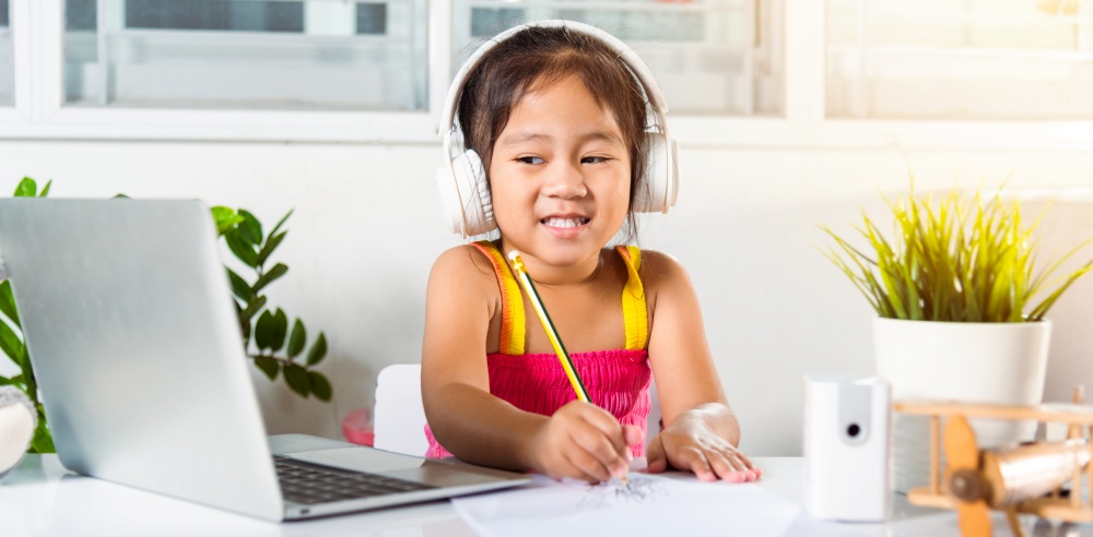 Asian child girl studying video conference distant education at home. Little kid preschool wear headphones sit at desk use laptop computer and communicates on Internet online video call with teacher