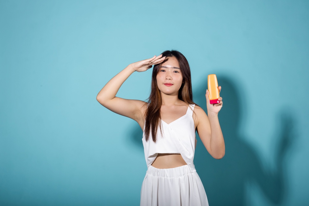 Beautiful brunette female hold cream sunblock bottle in studio shot isolated on blue background, Asian young woman holding body lotion on hand, tube of sunscreen, Beauty cosmetics facial treatment