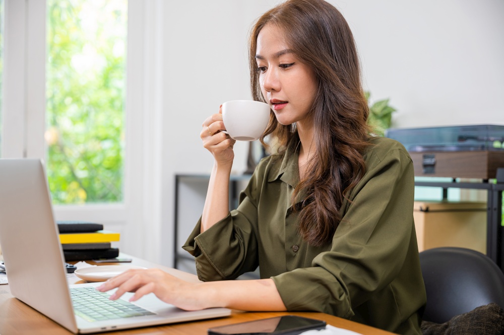Portrait of beautiful woman holding hot coffee mug cup on hand for drink while working online on laptop computer, smiling business woman drinks coffee sitting desk using laptop keyboard at morning