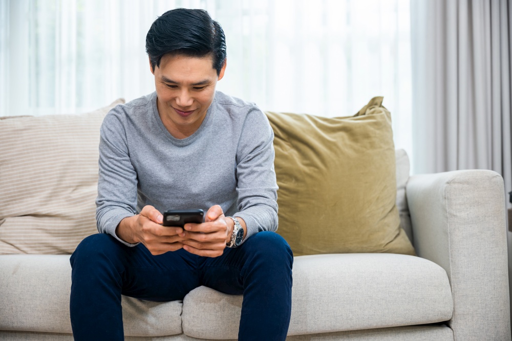 Asian older middle aged man relaxing sitting on sofa using smart mobile phone at home, Happy mature men smiling chatting with social media on smartphone in living room, holding device