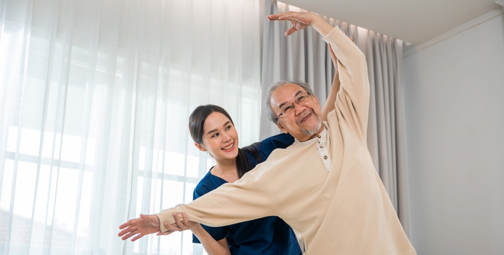 Rehabilitation of disabled people. Old senior man enjoys training with physiotherapist for outstretched arms at home, Asian physical therapist patient help elderly exercising arm stretch