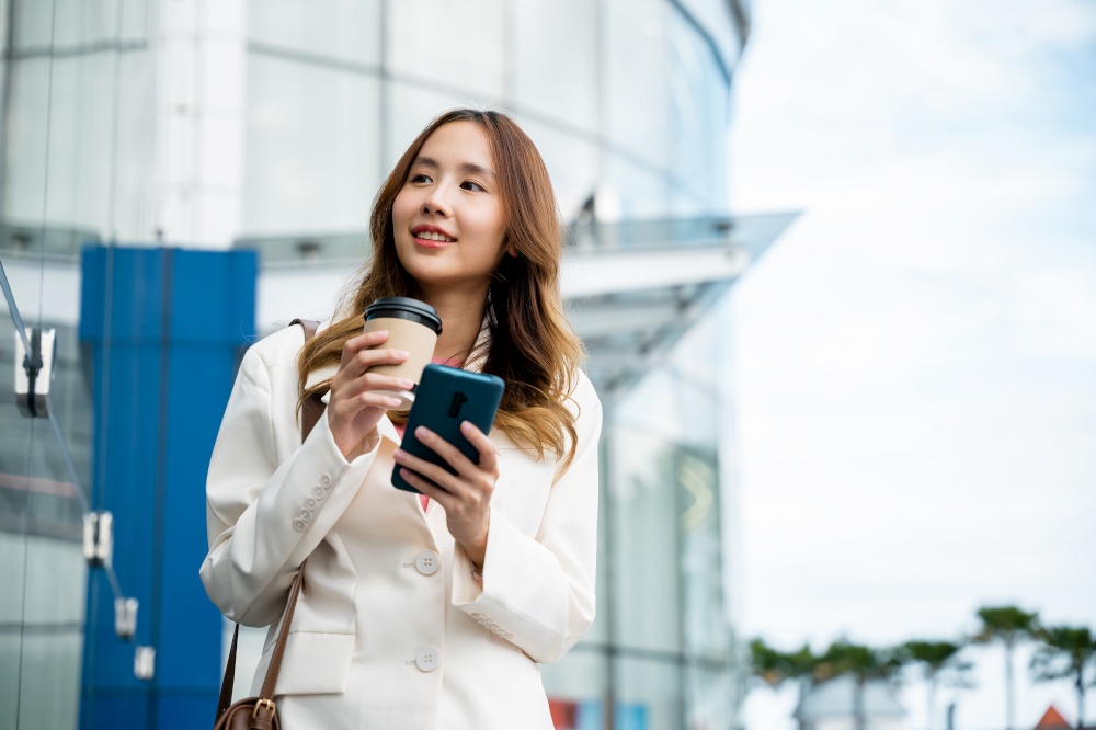 Asian businesswoman with smartphone and cup coffee standing street front building near office, Portrait woman smiling holding smart mobile phone with coffee take away going to work early in morning