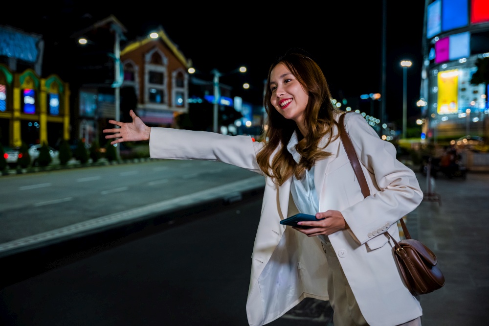 Beautiful woman smiling using smartphone application hailing with hand up calling cab after late work, Asian businesswoman standing hail waving hand taxi on road in busy city street at night