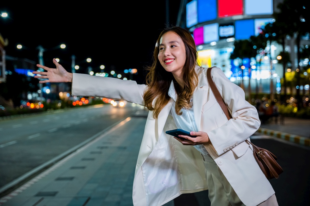 Beautiful woman smiling using smartphone application hailing with hand up calling cab after late work, Asian businesswoman standing hail waving hand taxi on road in busy city street at night