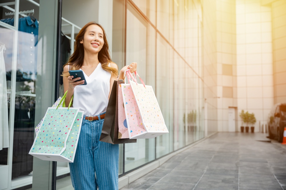 Happy Asian beautiful woman holding shopping bag and smartphone on her hands near the mall shop window, Smiling shopper customer girl lifestyle standing use mobile phone after shopping on city street