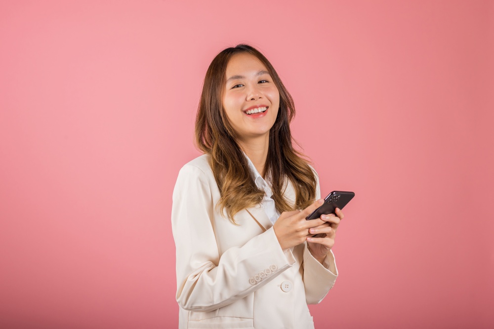 Happy Asian portrait beautiful cute young woman teen smiling excited hold smart mobile phone studio shot isolated on pink background, Female surprised and sms chatting internet online on smartphone