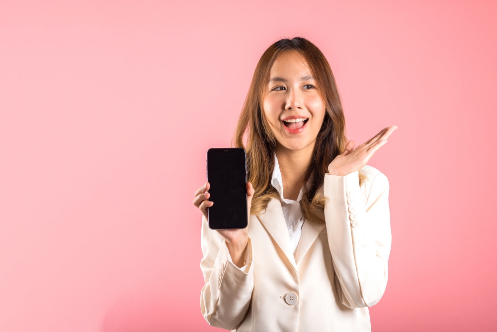 Happy Asian portrait beautiful cute young woman teen smiling excited holding blank screen mobile phone studio shot isolated on pink background, Thai female person showing smartphone empty screen space