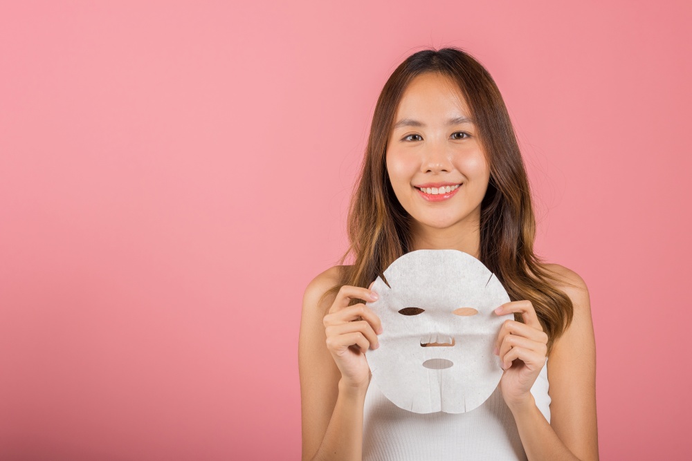 Healthcare spa. Portrait beautiful female smiling holds facial sheet mask, Studio shot isolated on blue background, Asian woman holding purifying mask face on hands, Skin care healthcare cosmetic
