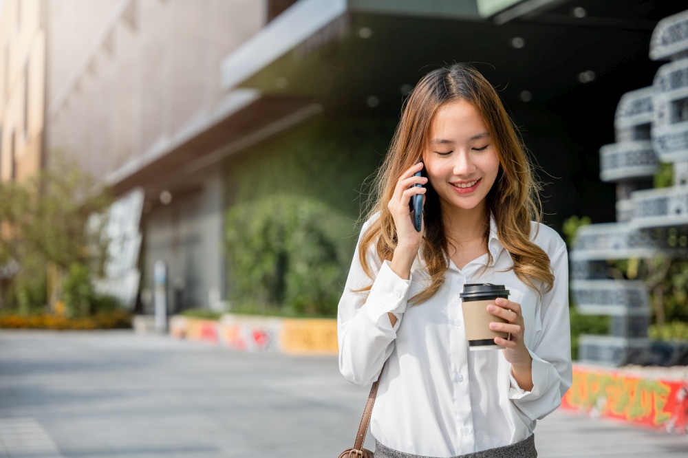Asian young businesswoman walking outdoors talking on cell smart mobile phone and holding coffee cup takeaway, lifestyle business woman hold paper cup and calling smartphone outside office