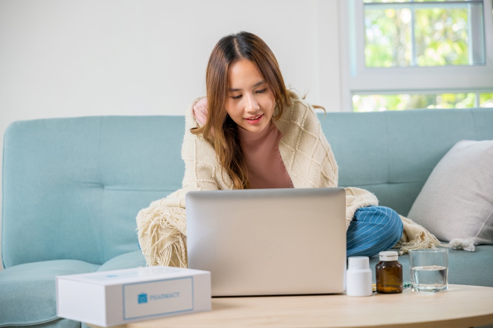 Sick young woman sit on sofa video call online with laptop consult with doctor at home, female covered blanket checking medications online, healthcare and technology