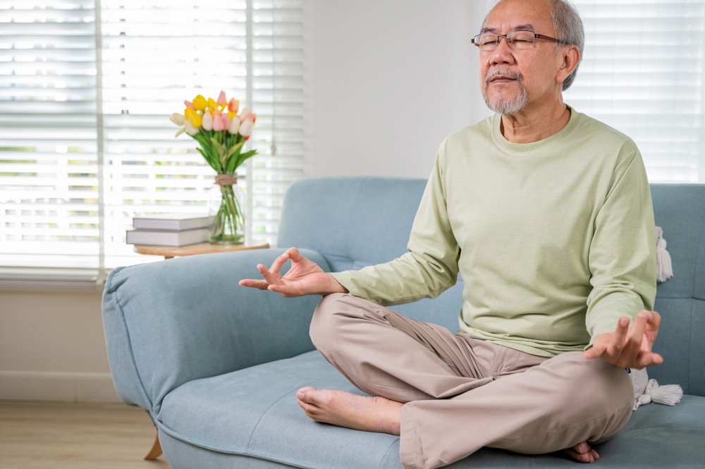 Asian old man practise yoga and meditation in lotus position and closed eyes, lifestyle senior man lotus pose doing yoga for mental balance breathing air relaxing on sofa at home, Healthy life habits