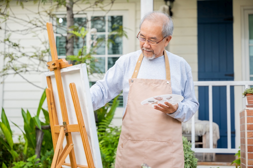 Lifestyle Asian senior old man painting picture artwork using brush and oil color on canvas, elderly people smile paint at his easel outside green nature background, Happy retirement artist activity