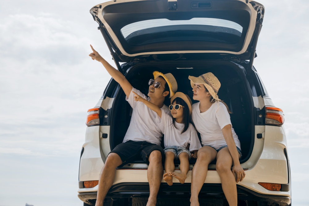 Tourism Day. Dad, mom and daughter enjoying road trip sitting on back car and pointing finger out blue sky, people having fun in summer vacation on beach, Family traveling in holiday at sea beach