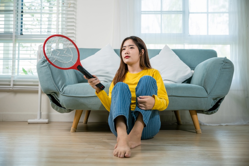 Asian young woman sitting floor using mosquito swatter or electric net racket, female killing mosquitoes she holding fly swatter like weapon in living room at home, Insect killer