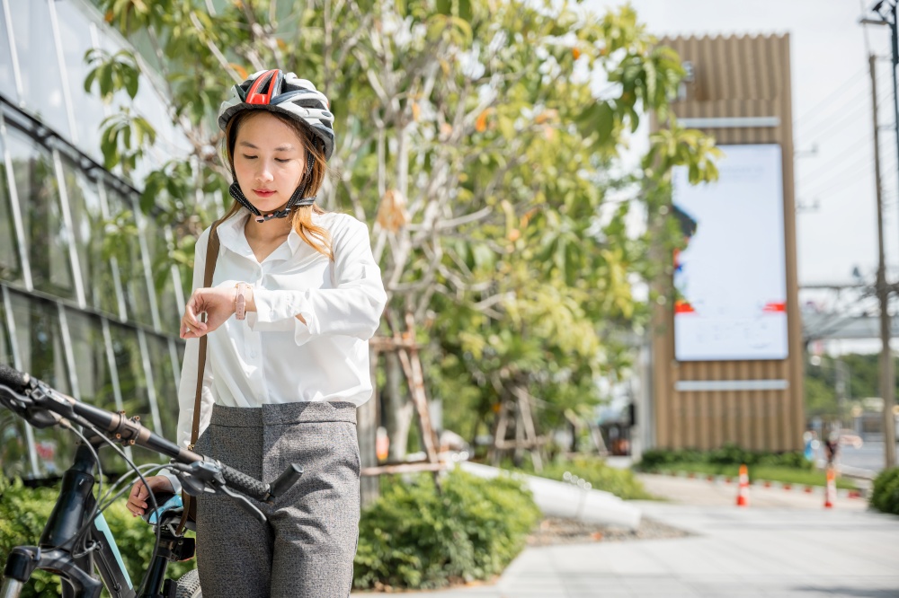 Asian young businesswoman standing with bicycle checking time on city street in morning before go to work at office, portrait smiling woman check watch, Commute to work, lifestyle biker