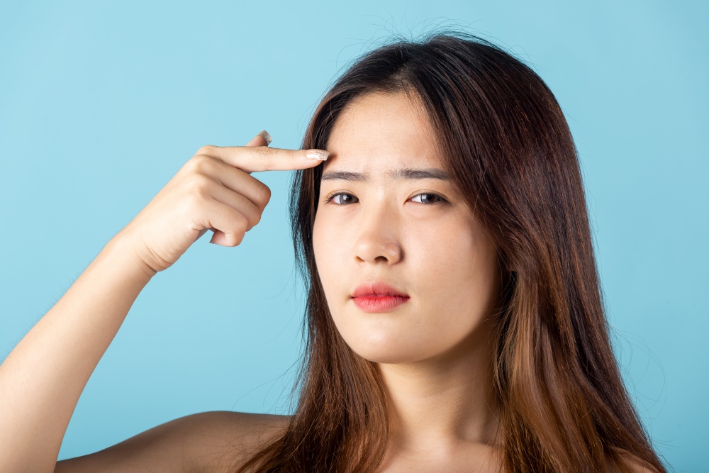 Beauty skin care concept. Asian young woman pointing eyebrow with her finger studio shot isolated on blue background, beautiful portrait female point face and forehead