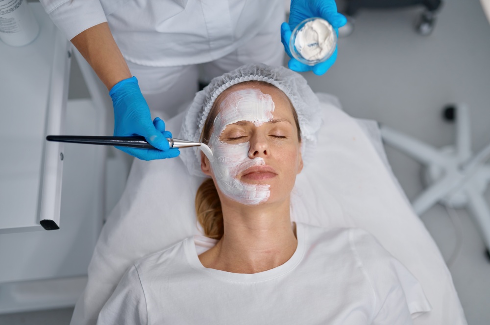 Cosmetologist applying mask on client face using brush. Professional cosmetology and beauty treatment in spa salon. Cosmetologist applying mask on client face in spa salon