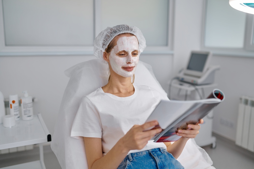 Woman client in clay face mask reading fashion magazine relaxing at cosmology office. Woman client in clay face mask relaxing at cosmology office