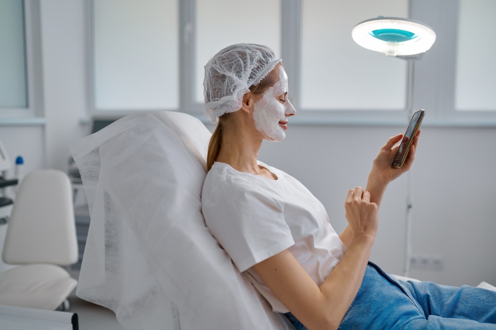 Woman client in clay face mask resting and using mobile phone for social network chatting. Cosmology service and beauty treatment concept. Woman client in clay face mask resting with mobile phone at cosmology office
