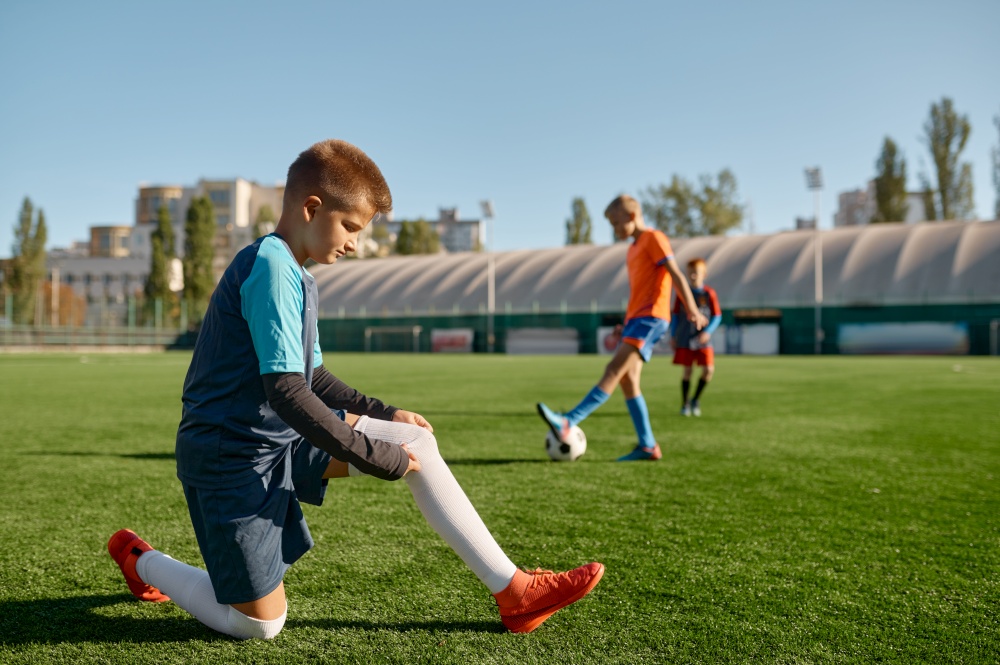 Active children playing football on green sport field outdoor. Focus on boy adjusting sock on foreground. Active children playing football on green sport field outdoor