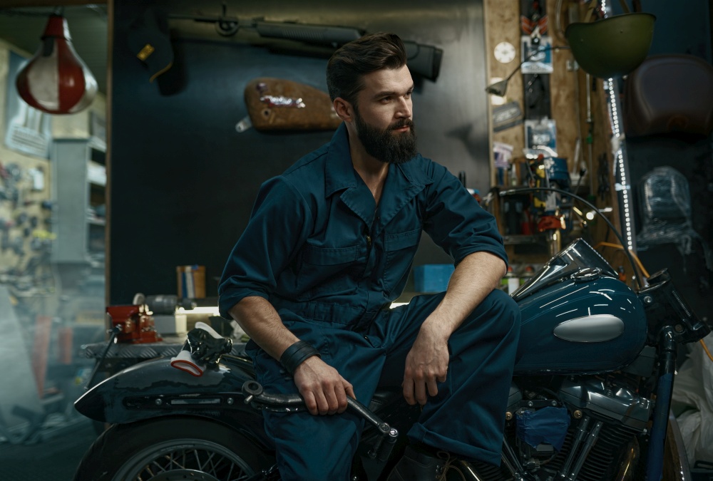 Portrait of confident man mechanic wearing overalls holding wrench in hand while sitting on repaired motorcycle. Workshop service concept. Portrait of confident man in overalls holding wrench while sitting on motorcycle