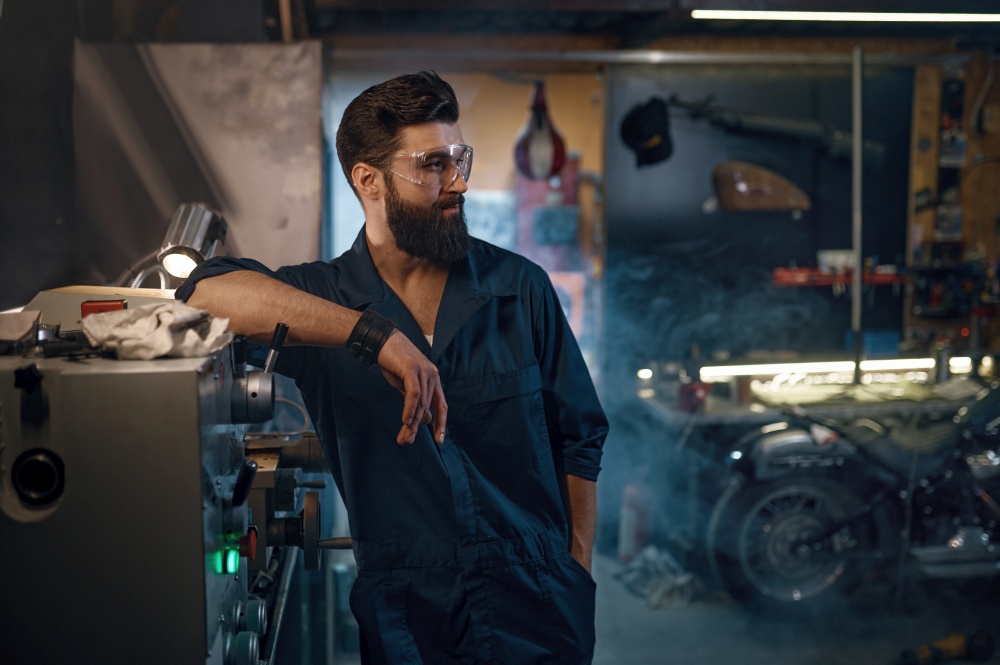 Portrait of confident handsome repairman in protective glasses and overalls standing in modern motorcycle workshop. Bike maintenance and repair professional service concept. Portrait of confident handsome repairman in protective glasses and overalls