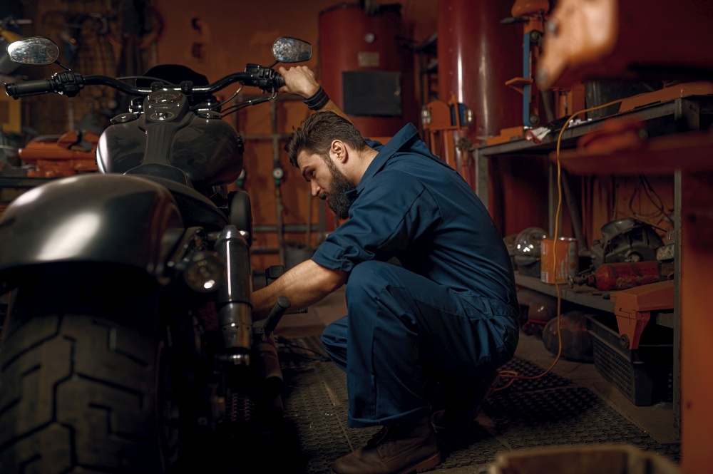 Mechanic working with motorcycle engine in workshop. Technician in blue workwear sitting nearby vehicle. Mechanic working with motorcycle engine in workshop while sitting nearby vehicle