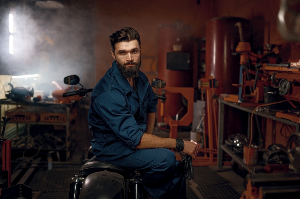 Portrait of tired but happy workman sitting on repaired motorcycle in workshop. Young adult bearded repairman looking at camera. Portrait of tired but happy workman sitting on repaired motorcycle in workshop