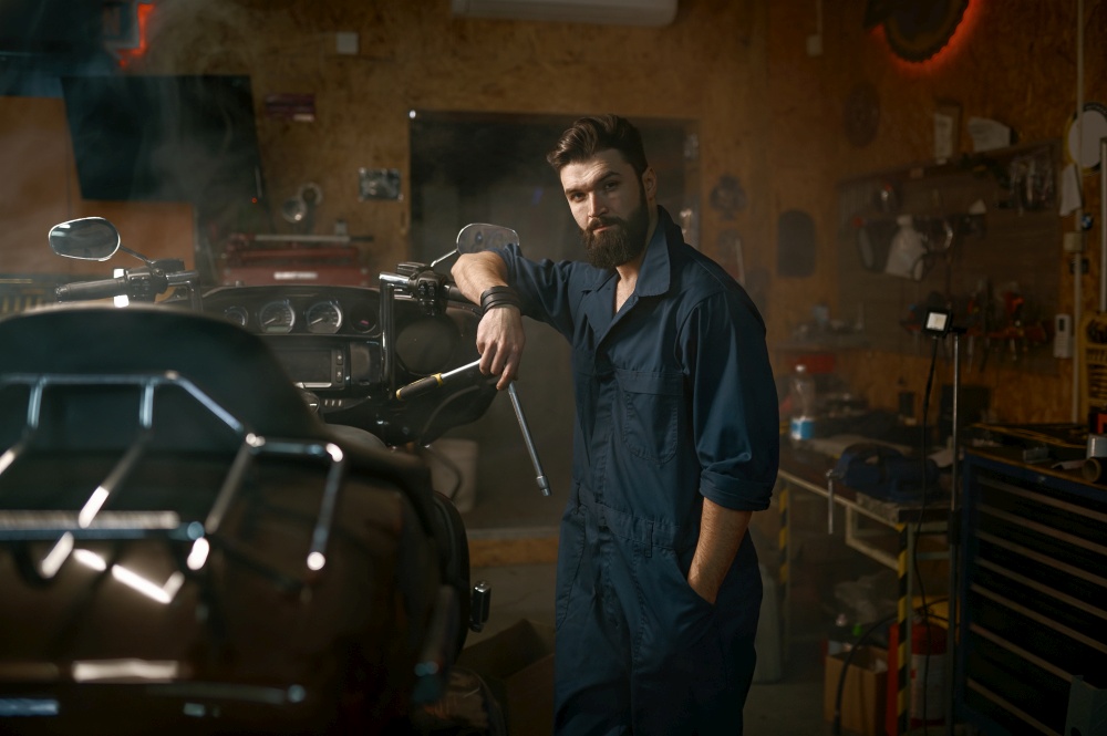 Handsome brutal repairman in work wear looking at camera portrait. Bearded young workman standing nearby motorcycle and holding wrench over smoky dark repair garage workplace. Handsome brutal repairman in work wear looking at camera portrait